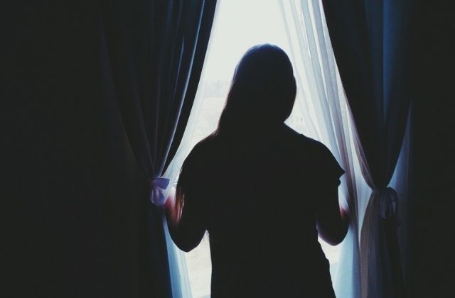 A dark-lit image of a woman with her back to the camera as she looks out the window