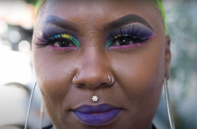 Black woman with very close cropped blonde hair, and colorful eye make up (green, yellow, purple) as well as several nose rings and purple lipstick