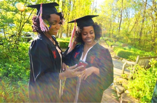 Two Black women dressed in black graduation caps and gowns look at an image on a cell phone