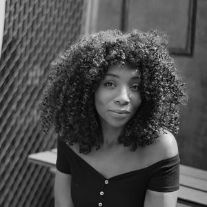 Black woman with black dress and brown curly hair sitting.