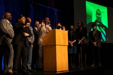About 20 people onstage with a image of George Floyd on a screen behind them. There is a podium and microphone. Press Conference