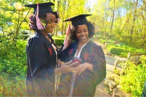 Two Black women dressed in black graduation caps and gowns look at an image on a cell phone